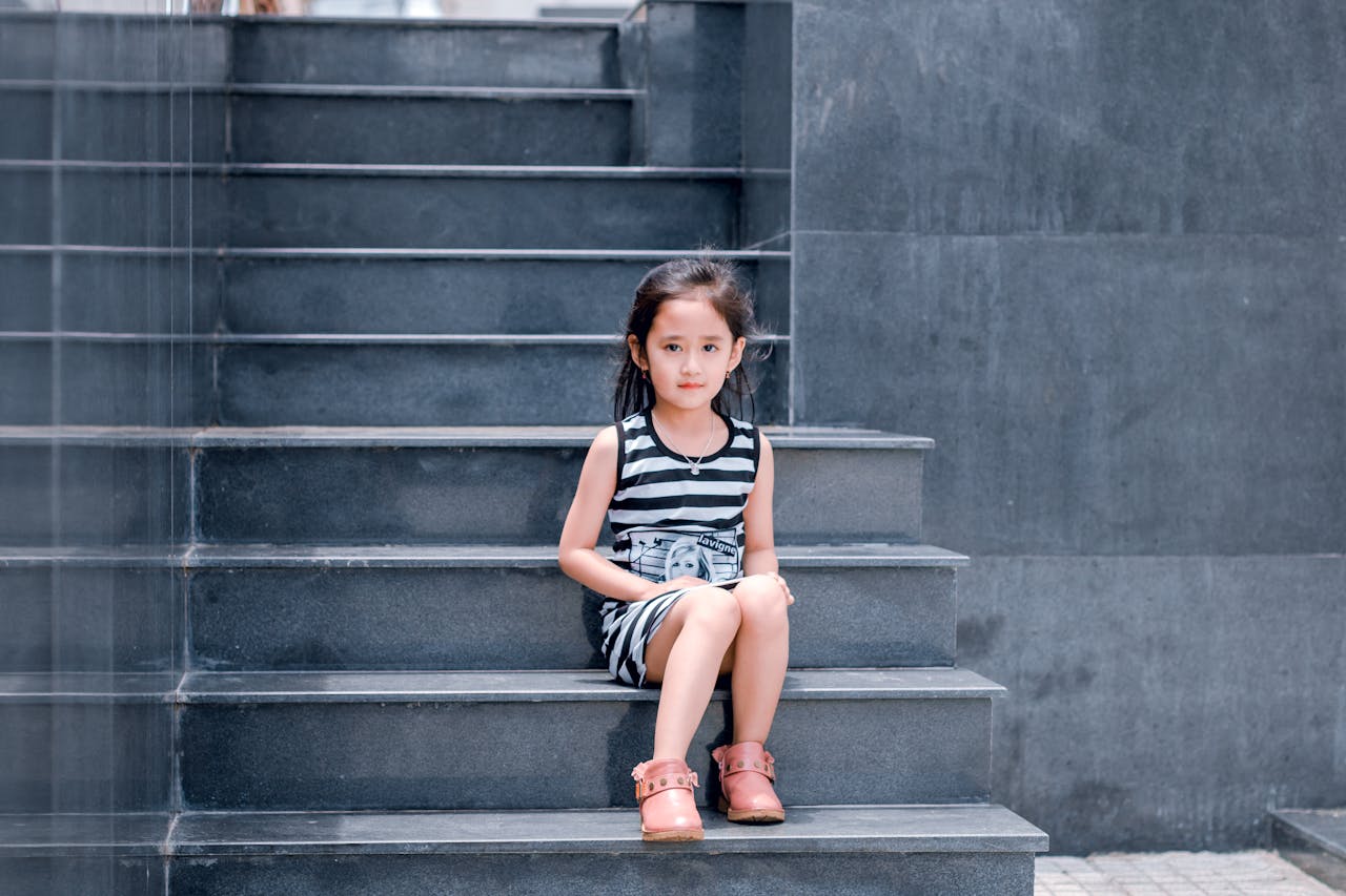A young girl in a striped dress sits on a sleek grey staircase, looking into the camera.