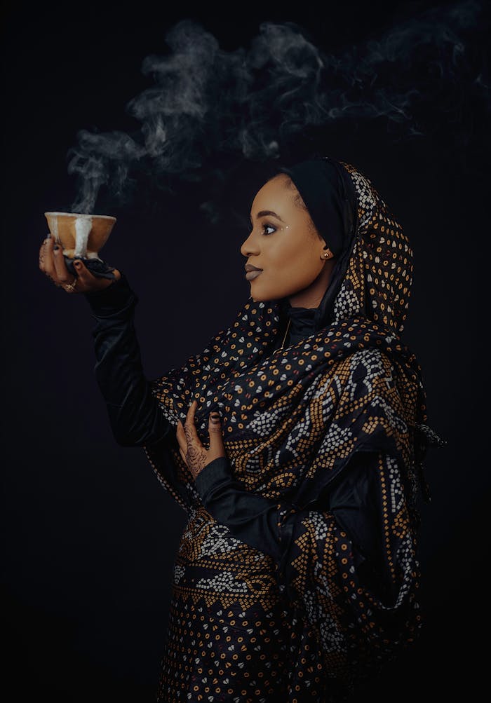 Portrait of a beautiful African woman in Nigerian attire holding a steaming cup against a dark backdrop.