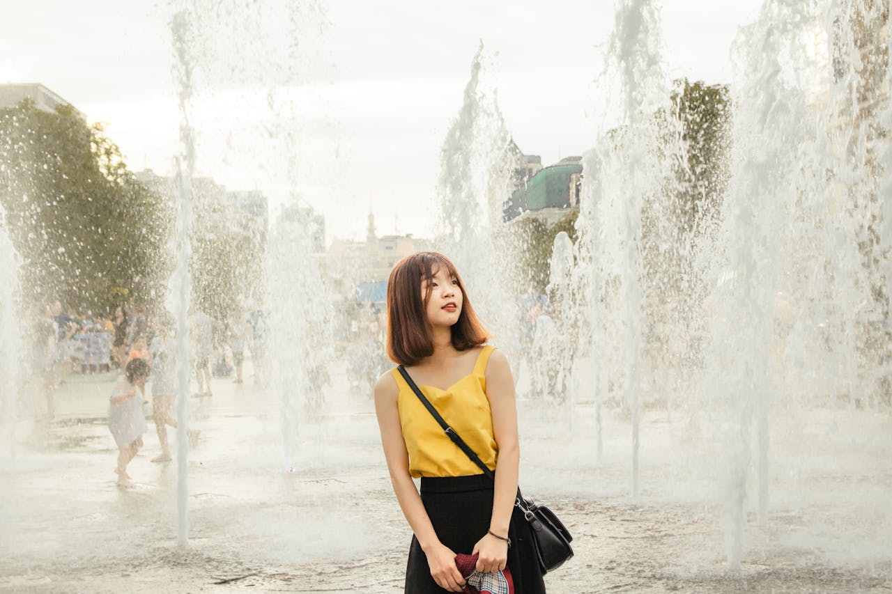 A young woman in a yellow top stands near water fountains outdoors, showcasing a serene and fashionable scene.